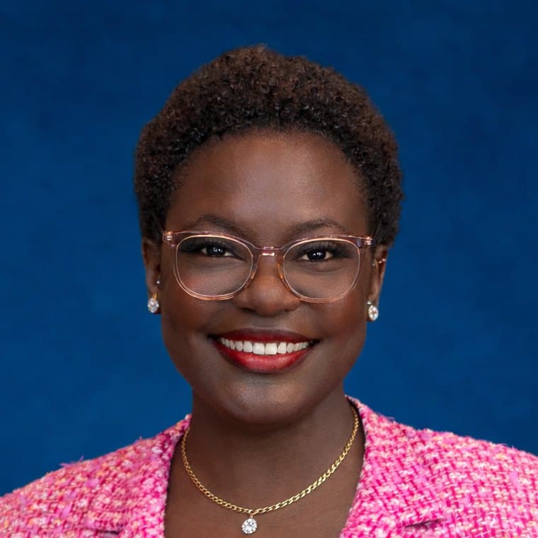 Dr. Raven Baxter smiling in glasses and a pink tweed jacket, a studio headshot on cobalt