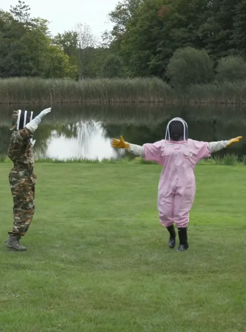 Dr. Raven Baxter in a pink beekeeping suit and a beekeeper in a camouflage suit holding their arms out on a lawn by a pond, from Nerdy Jobs Episode 2
